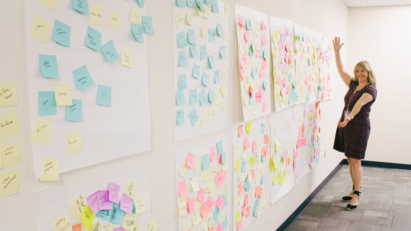 Justine beside a long wall covered in colorful sticky notes from a workshop, arm raised in celebration